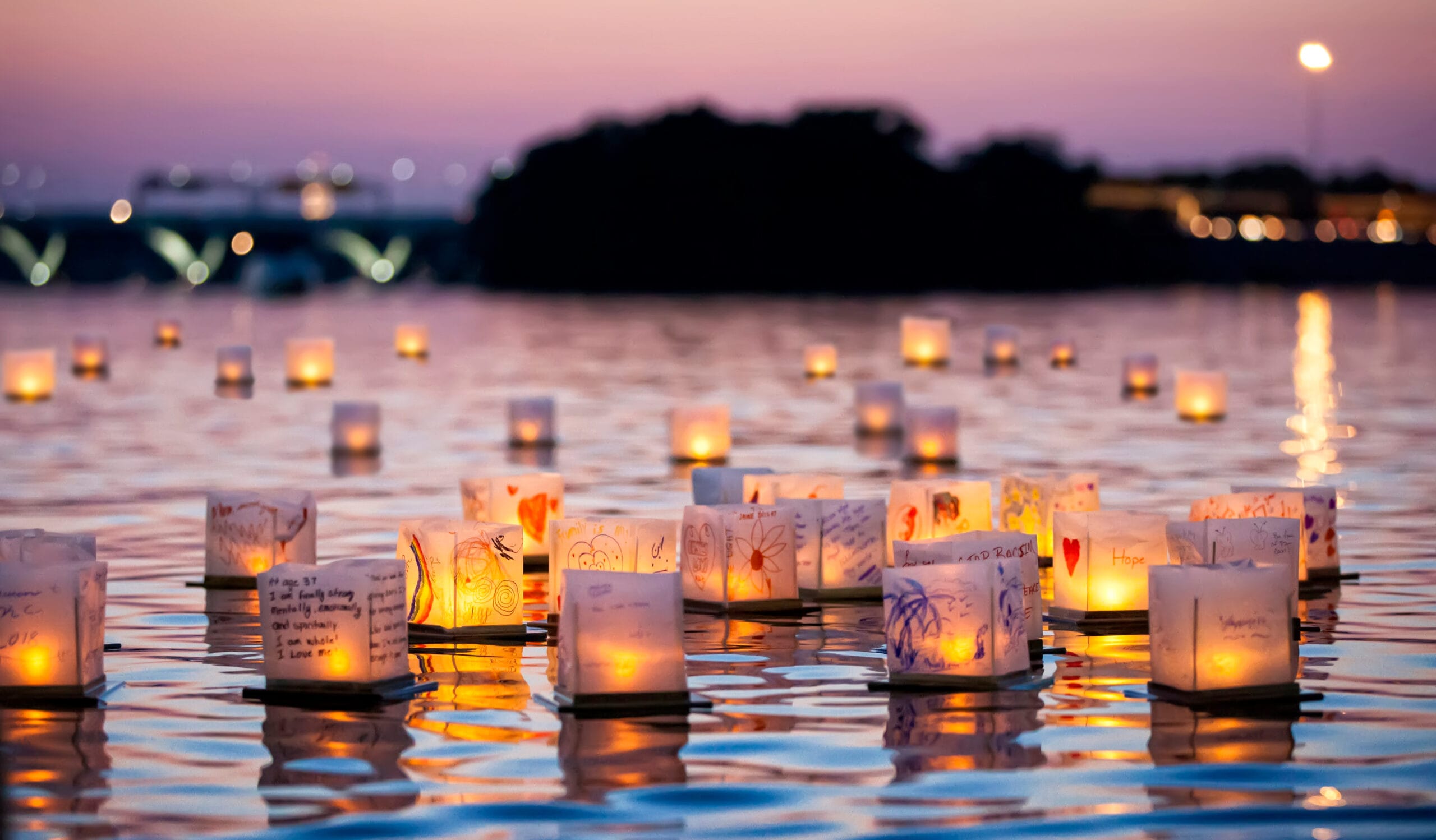 Square lanterns covered in illustrations float on blue and pink water at sunset.